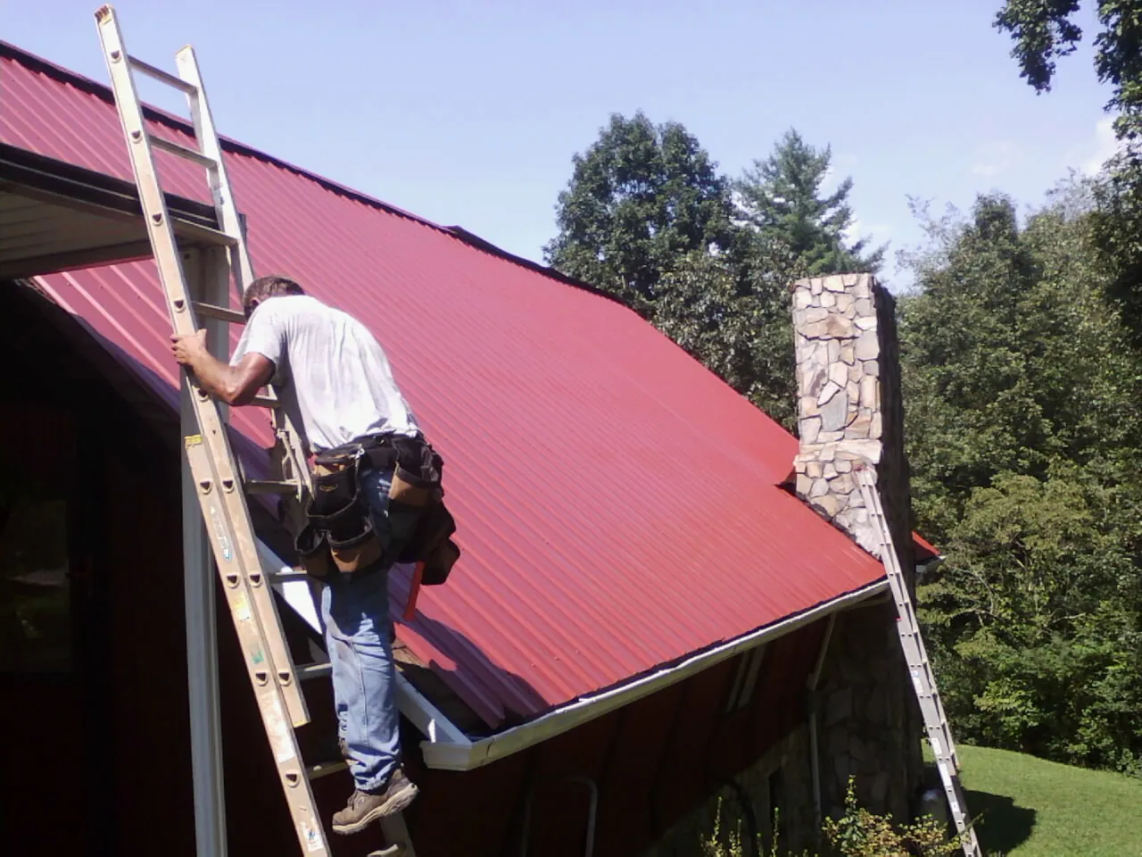 Roofer on a ladder beside a red metal roof and stone chimney — Soffit & Fascia Repair by Roofing Brothers in Mound Valley
